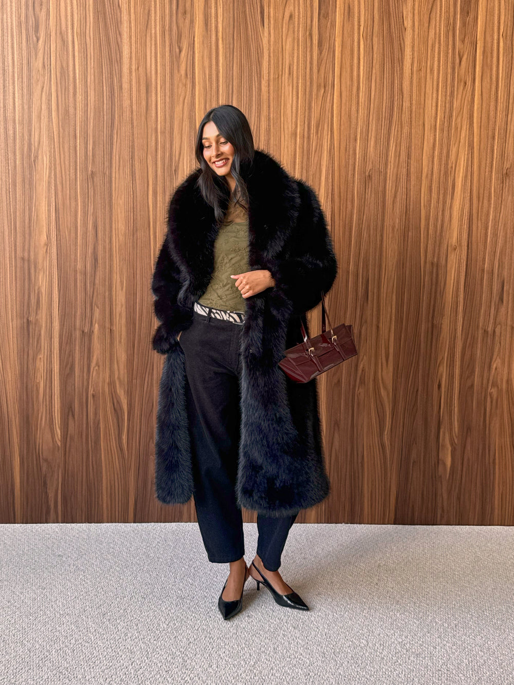 Long dark brown faux fur coat over an olive green top and dark trousers.
